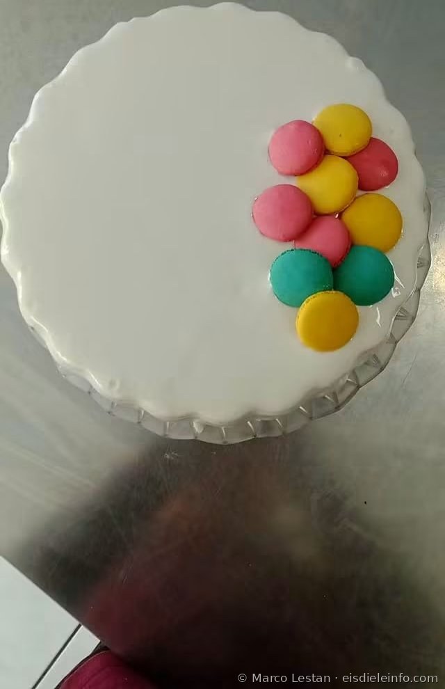 Eistorte mit weißer Glasur und bunten Macarons in Rosa, Gelb und Türkis