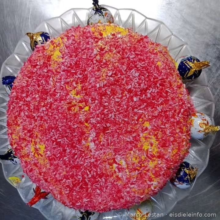 Eistorte mit rosa-rotem Granulat-Überzug, umgeben von verpackten Pralinen