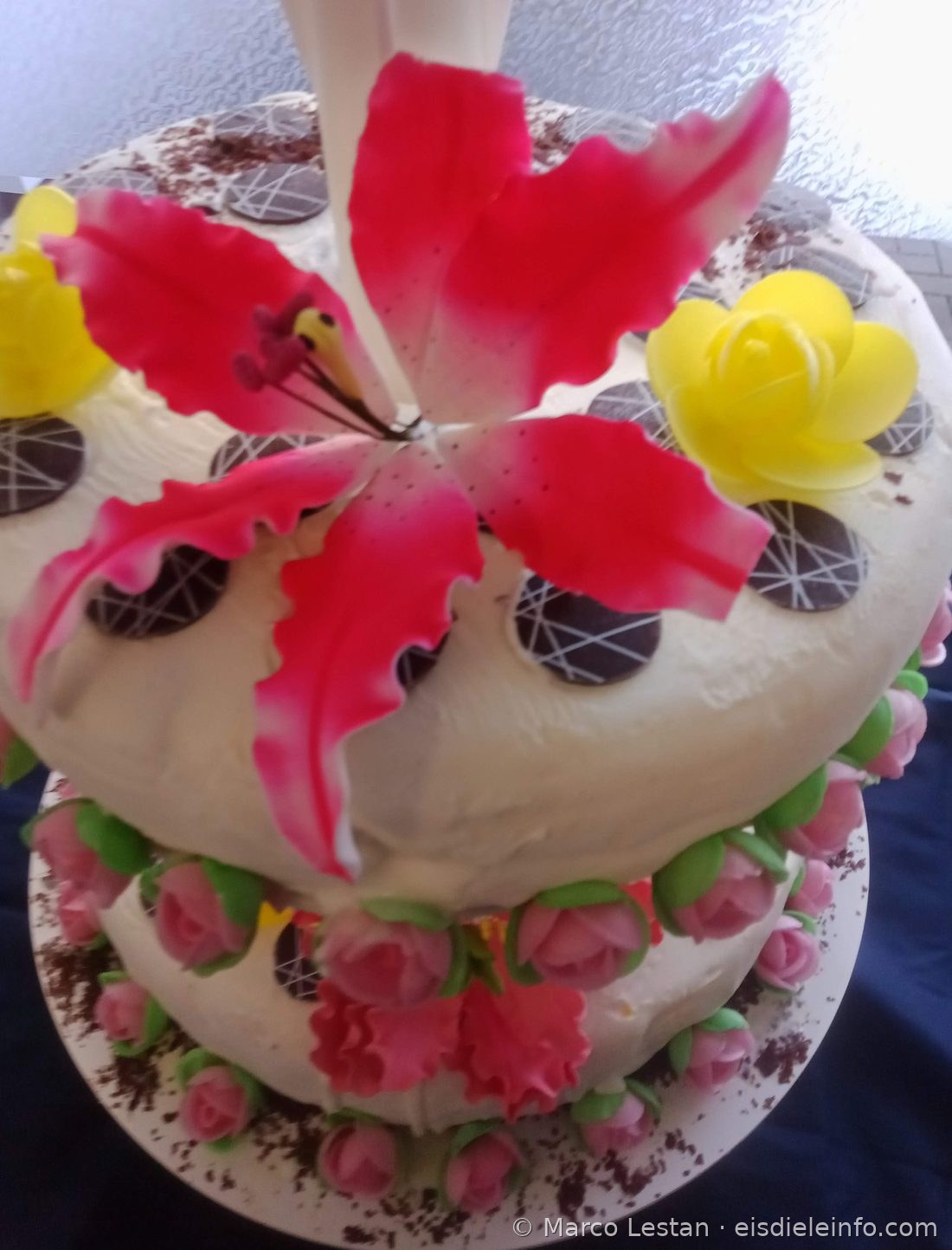 Rosa Zuckerpaste-Lilie auf weißer Eistorte mit gelben Oblatenrosen und Schokoladenscheiben