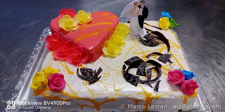 Rechteckige Torte mit weißer Glasur, rosa Herz, gelben und roten Rosen, Schokoladenspänen und Brautpaar